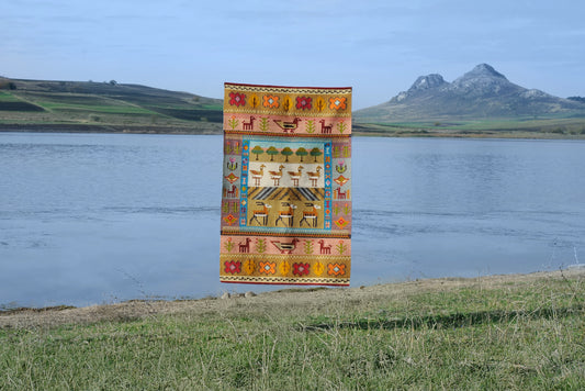 Colorful tapestry with geometric patterns of Gazelles and pistachio trees displayed outdoors by a body of water and mountains.